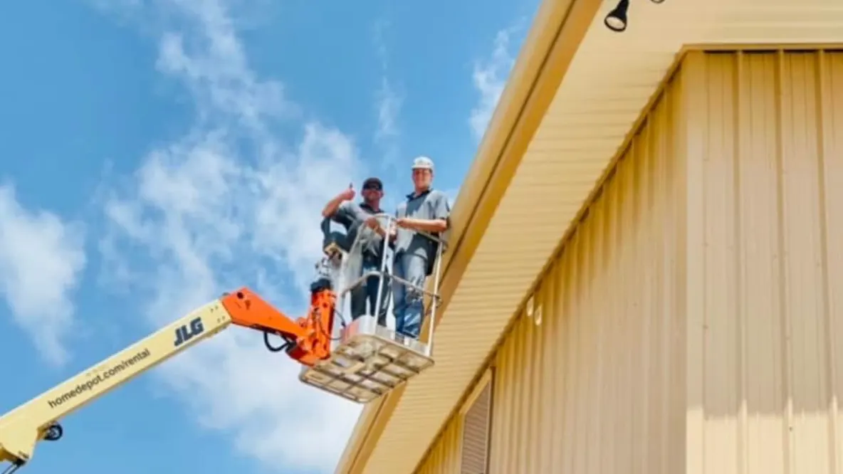 Two PSG crew members on a lift at the eave of a tan metal building, one giving a thumbs up