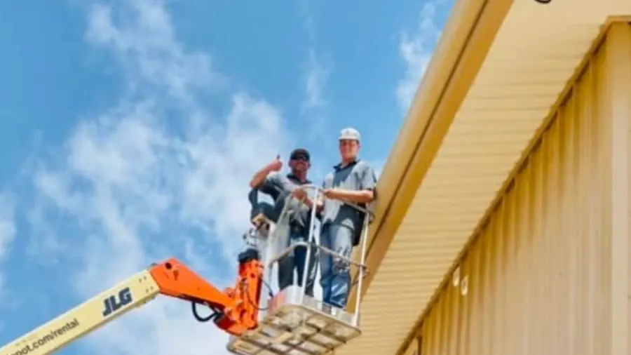 Two PSG crew members on a lift at the eave of a tan metal building, one giving a thumbs up
