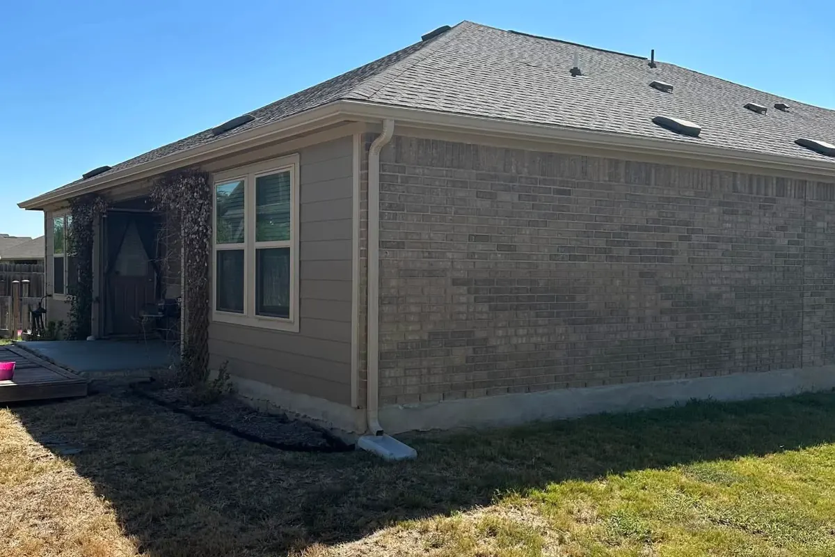 A brick and siding house with a new gutter along the eave and a tan downspout at the corner emptying onto a white splash block