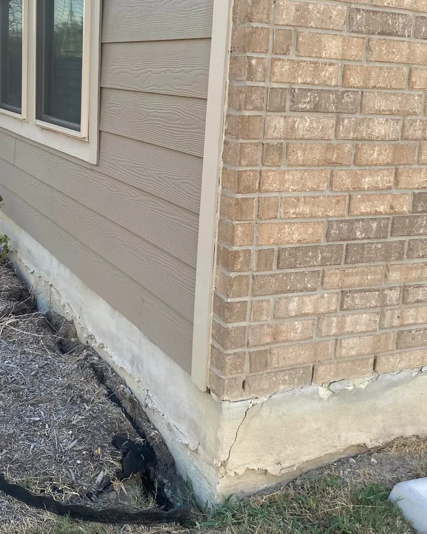 The corner of a brick and siding house where water has washed the soil and mulch away from the slab, leaving the concrete edge exposed, cracked and crumbling, with the garden edging pulled loose
